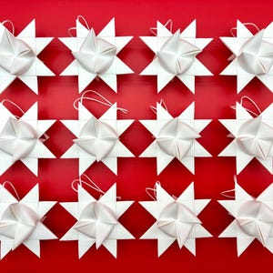 Może przedstawiać: Układ dwunastu białych, gwiazdkowych ozd&oacute;b świątecznych ze złożonym środkiem wstążki, na tle żywego czerwonego tła. Każda gwiazda ma ostre krawędzie i pętlę do zawieszenia. Świąteczna dekoracja świąteczna.