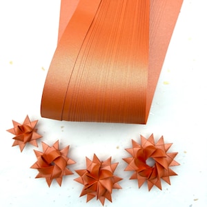 May include: Close-up of orange paper quilling supplies. Several star-shaped quilled paper ornaments are arranged near a stack of orange paper strips. The paper is a solid, matte orange color, and the background is white.