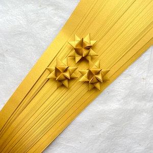 May include: A close-up shot features three golden star ornaments resting on a diagonal arrangement of gold paper strips. The stars are made of folded paper, and the background is a textured white surface.