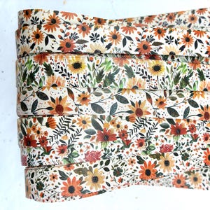May include: A stack of floral patterned rolls. The rolls feature a repeating pattern of orange, yellow, and red sunflowers, leaves, and other flowers on a cream background. The floral design is detailed and colorful.