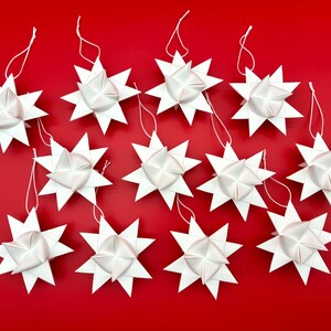 Może przedstawiać: Kolekcja białych ozd&oacute;b w kształcie gwiazd z ostrymi krawędziami i centralnym, złożonym wzorem. Każda ozdoba ma sznurek do zawieszenia i jest umieszczona na żywym czerwonym tle, idealna do dekoracji świątecznych.