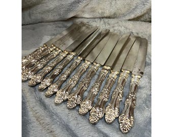 May include: A collection of silver-colored butter knives. Each knife has a decorative handle with floral detailing. The blades are rectangular and appear to be made of stainless steel. The knives are arranged on a soft, gray surface.
