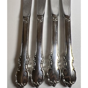 May include: Four silver butter knives with ornate, floral-style handles. The blades are smooth and reflective, and the handles feature a detailed, embossed design. The knives are arranged vertically, side by side.