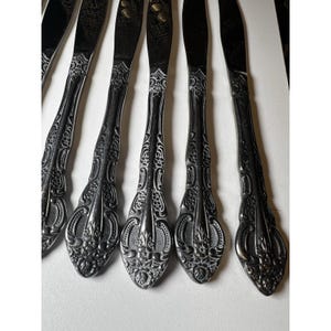 May include: Close-up of several ornate silver knives. The handles feature a detailed floral and scroll design, with a pointed tip. The blades are dark and reflect light. The knives are arranged in a row, suggesting a set of silverware.