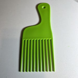 May include: A bright green Afro pick comb with a handle and a series of long, evenly spaced tines. The comb is made of plastic and has a hole at the top of the handle.