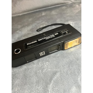 May include: Black Kodak Ektralite 10 camera with a built-in flash. The camera has the Kodak logo and the text "Ektralite 10 camera" printed on the top. It has a small viewfinder and a lens on the front.