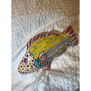 May include: A ceramic fish figurine with a colorful, hand-painted design. The fish features a yellow and green checkered pattern on its back, red and white striped fins, and a body adorned with red, yellow, and blue accents. The figurine is set against a white, textured background.
