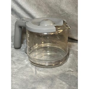 May include: A clear glass coffee carafe with a gray plastic handle, lid, and spout. The carafe has measurement markings in ounces and a capacity of up to 25 ounces. The handle is on the left side of the carafe.