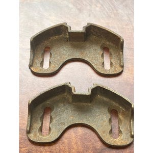 May include: Two antique, weathered metal brackets with a curved bottom and a rectangular top. Each bracket has two elongated, vertical slots for mounting. The brackets have a dark, aged appearance.
