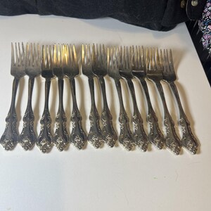 May include: A collection of twelve silver forks arranged in a row. The forks have ornate, detailed handles with a floral design. The tines of the forks are visible, and the overall appearance is vintage or antique.