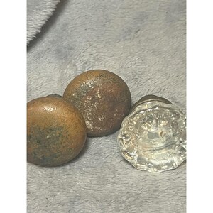 May include: Three antique door knobs. Two are round and made of aged, tarnished metal with a bronze hue. The third is a clear, faceted glass knob with a decorative design. The knobs are resting on a soft, gray surface.