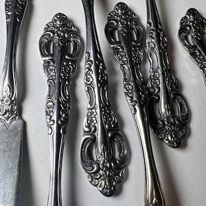 May include: Close-up of ornate silverware with a floral design. The silver utensils include forks, knives, and spoons. The handles have a detailed, embossed pattern. The silverware is arranged on a white surface.