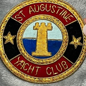 May include: A round emblem with the text "ST AUGUSTINE YACHT CLUB" in gold lettering. The emblem features a central design of a gold tower on a blue and light blue background, surrounded by a red border and gold stars.