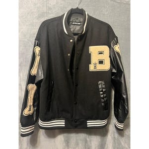 May include: Black varsity jacket with gold accents. Features a large gold "B" patch and bone-shaped designs on the sleeves. The jacket has black leather sleeves and a white and black striped hem and cuffs. The collar is white.