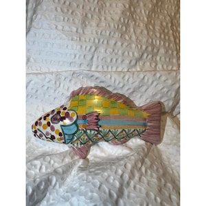May include: A colorful ceramic fish figurine with a whimsical design. The fish features a checkered yellow and green pattern on its back, with pink fins and a tail. The head is decorated with polka dots and other colorful details.