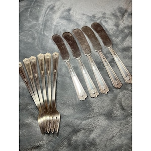 May include: A collection of vintage silverware, including butter knives and forks. The silverware has a silver finish with ornate detailing on the handles. The butter knives have a rounded blade. The forks have three tines.