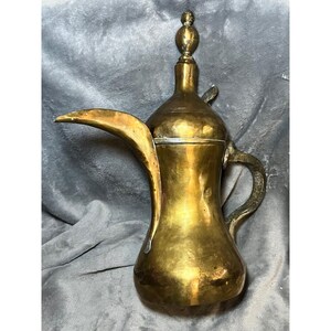 May include: A vintage brass coffee pot with a long, curved spout and a decorative handle. The pot has a bulbous body and a tall, slender neck with a small lid. The overall design is ornate and traditional, with a polished finish.