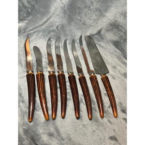 May include: A set of eight vintage knives with stainless steel blades and brown, faux-antler handles. The set includes various knife types, including a carving knife and steak knives, arranged on a gray surface.
