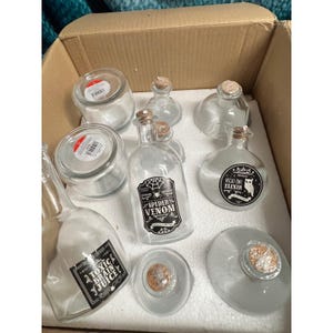 May include: A collection of clear glass potion bottles and jars with cork stoppers. Some bottles have labels reading "Spider Venom", "Toxic Brain Juice", and "Night Owl Elixir". The items are arranged inside a cardboard box.