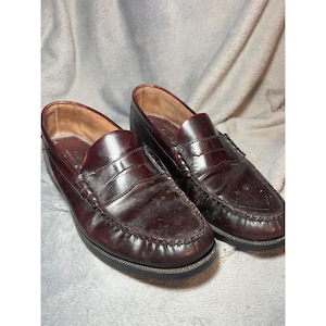 May include: A pair of burgundy leather penny loafers. The shoes have a classic design with a rounded toe, a strap across the vamp, and a stitched sole. The leather appears worn, with some creases and scuffs.