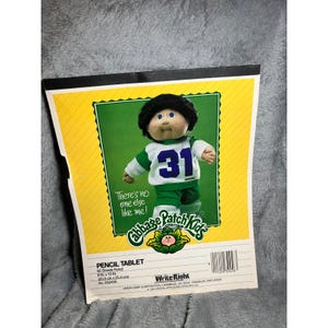 May include: A Cabbage Patch Kids pencil tablet with a picture of a doll wearing a green and white jersey with the number 31. The tablet has 60 ruled sheets and measures 8 in x 10 in.