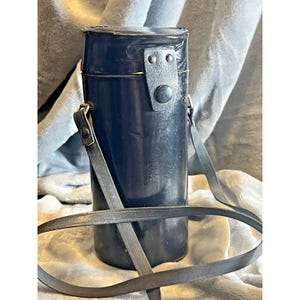 May include: A cylindrical, dark blue leather case with a shoulder strap. The case has a flap closure secured by a button. The strap is attached to the case with metal hardware. The case appears to be designed to hold a camera lens or other similar item.