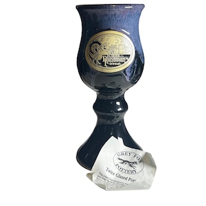 May include: A dark blue and black ceramic goblet with a flared top and a pedestal base. The goblet has a gold emblem that reads "Florida Renaissance Festival 2000". A paper tag from Grey Fox Pottery is attached.
