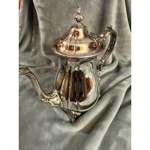 May include: A silver-colored teapot with a decorative lid and handle. The teapot has a curved spout and sits on three small feet. The lid is topped with a decorative finial. The teapot is highly reflective, showing the surrounding environment.