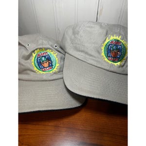May include: Two beige baseball caps featuring an embroidered circular patch on the front. The patch displays a brown coffee mug with a globe inside, surrounded by a yellow sunburst design and the text Planet Java in red and blue lettering.