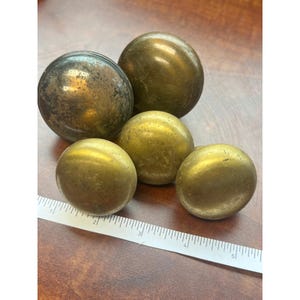 May include: A collection of five antique brass spheres of varying sizes. The spheres have a tarnished, aged appearance, with colors ranging from dark bronze to golden yellow. A ruler is placed near the spheres for scale.
