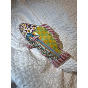 May include: A decorative ceramic fish figurine with a colorful design. The fish features a yellow and green checkered pattern on its body, with red and white striped fins. The head is adorned with a pattern of red, yellow, and brown dots.