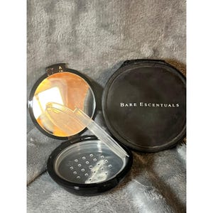 May include: Open black compact with a mirror, a sifter, and a powder compartment. The lid of the compact is open, revealing the mirror. The sifter is clear plastic. The brand name "Bare Escentuals" is printed on the lid.