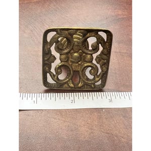 May include: Ornate brass square decorative hardware with an openwork design. The hardware features a symmetrical pattern of swirls and floral elements. The piece is approximately 2.5 inches wide and is set against a brown surface.