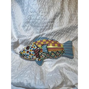 May include: A colorful ceramic fish-shaped decorative piece. The fish features a vibrant design with polka dots, checkered patterns, and geometric shapes in various colors, including blue, yellow, red, and green. The fins are blue.