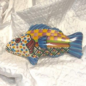 May include: A colorful ceramic fish figurine with a whimsical design. The fish features a checkered pattern in yellow and purple, with blue, red, and green accents. The body is decorated with polka dots, and the fins are blue. The figurine is approximately 8 inches long.