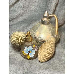 May include: A collection of vintage perfume bottles. One bottle is clear with a blue floral design. Another is a large, silver-colored bottle with a gold-colored atomizer. A third bottle is a gold-colored sphere.