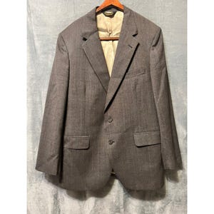 May include: A gray blazer with a two-button closure, notched lapels, and two front pockets. The blazer is hanging on a wooden hanger against a light blue background. The interior lining is a light tan color.