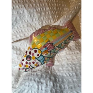 May include: A colorful ceramic fish figurine with a whimsical design. The fish features a white head with polka dots, a yellow and green checkered body, and pink fins. Decorative item.