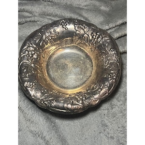 May include: A small, round, silver-colored bowl with a decorative grape and leaf pattern around the rim. The interior of the bowl has a gold-colored finish. The bowl is likely made of metal and is suitable for serving small portions of food or as a decorative item.