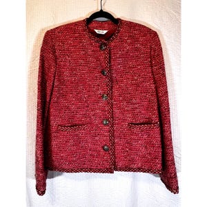 May include: A red tweed jacket with a round collar and four decorative buttons. The jacket has two front pockets and is trimmed with a gold and red border. The jacket is hanging on a black hanger.
