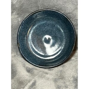May include: A small, round, speckled blue ceramic bowl with a dark rim. The bowl has a smooth interior and a slightly textured exterior. The bowl is likely used for serving food or as a decorative item.