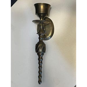 May include: A vintage brass wall sconce with a decorative design. The sconce features a cup-shaped holder, a small circular shelf, and a long, ornate stem with a spiral and beaded pattern. The sconce is designed to hold a candle and is mounted on a circular backplate.