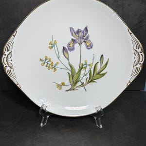 May include: White serving platter with a gold rim and handles. The platter features a floral design with a purple iris, yellow flowers, and green leaves. The platter is square with rounded edges.