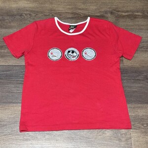 May include: Red t-shirt with a white-trimmed neckline and short sleeves. The shirt features three circular embroidered patches with a Mickey Mouse design and other graphics.
