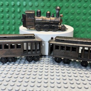 May include: A set of three bronze-toned metal toy train cars, including one steam locomotive and two San Francisco cable car style passenger carriages, each featuring detailed wheels, open window frames, and embossed lettering on the sides.