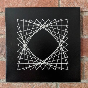 May include: A black canvas with a white string art design of a geometric shape. The design is made up of a series of triangles that create a diamond shape in the center.