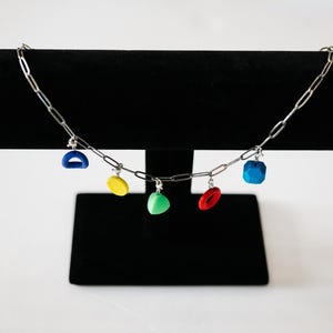May include: A silver chain necklace with five colorful charms: a blue arch, a yellow circle, a green triangle, a red oval, and a blue square. The necklace is displayed on a black velvet stand against a white background.