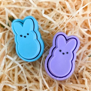 May include: Two bunny-shaped cookie cutters in light blue and lavender. Each cutter has a black outline and three small dots for eyes. The cutters are resting on a bed of light brown wood shavings.