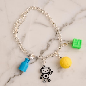 May include: Silver charm bracelet with a chain link design. The bracelet features several colorful charms, including a blue bottle, a black and white robot, a yellow ball, and a green square. The bracelet is displayed on a marble surface.
