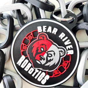 Peut inclure: Un patch circulaire noir et rouge avec le texte "Bear River Robotics" en lettres blanches. Le patch représente un ours stylisé avec un visage rouge et gris.